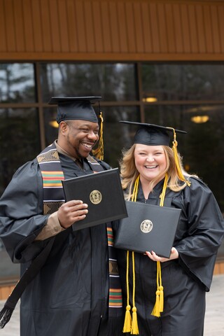 two graduates outside in cap and gown with their diplomas