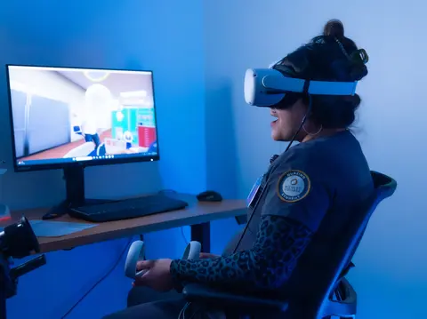 Person using VR headset and controllers, seated by a computer monitor.